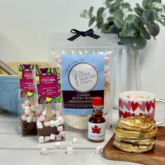 Gourmet pancake mix with two hot chocolate stirrers, mini bottle of maple syrup, stack of pancakes and mug of hot chocolate and marshmallows.