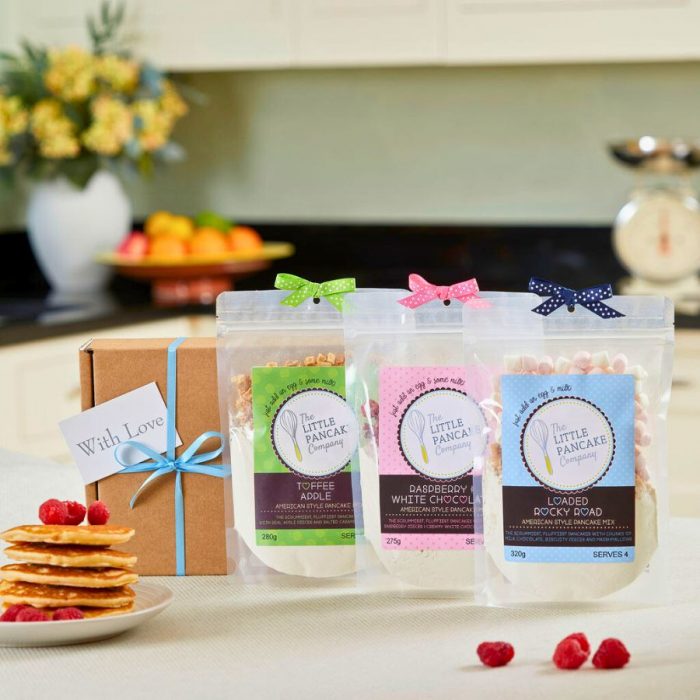 Three pancake mixes, a stack of pancakes, a gift box in a kitchen setting